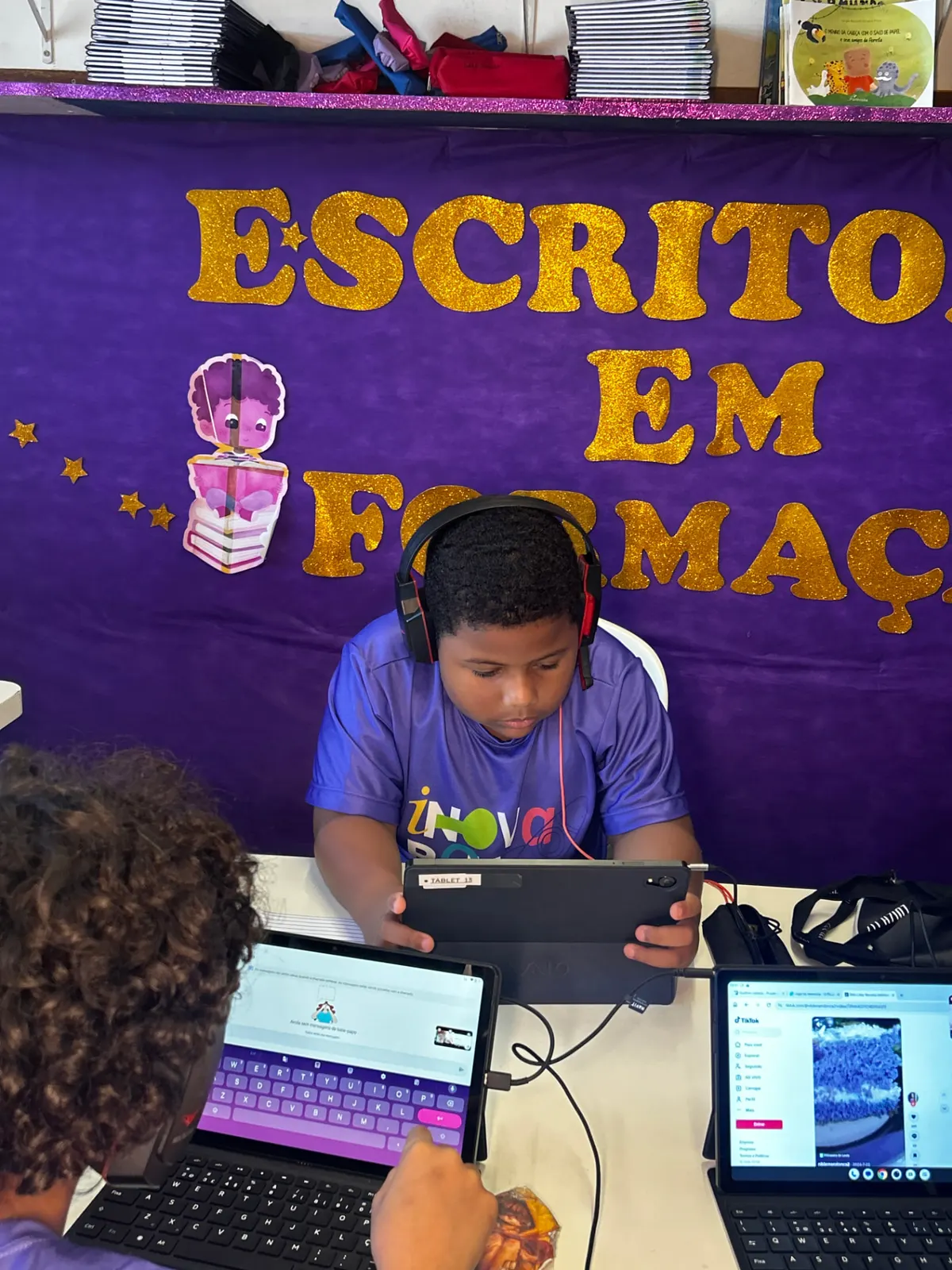 Aluno em aula online com tablet e fone no painel Escritores em Formação